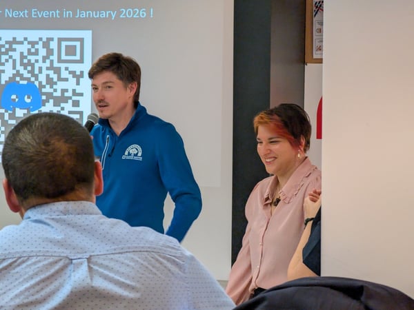 A candid photograph taken indoors at a technical or professional event, showing a man speaking into a microphone next to a smiling woman, with a presentation slide visible behind them featuring a QR code and the text, "Next Event in January 2026 !"