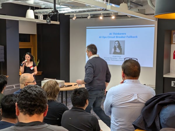 A photograph capturing an indoor tech presentation where a woman is speaking into a microphone to a seated audience, with a slide titled 'AI Ops Circuit Breaker Fallback' and featuring the speaker's name, Angie Ariza, projected behind her.