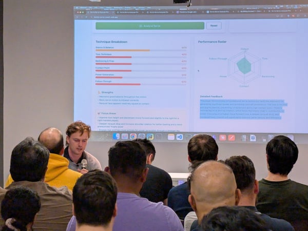 A photograph taken during an indoor presentation where a speaker, holding a microphone, addresses an audience while displaying a tennis serve analysis application on a large projected screen.