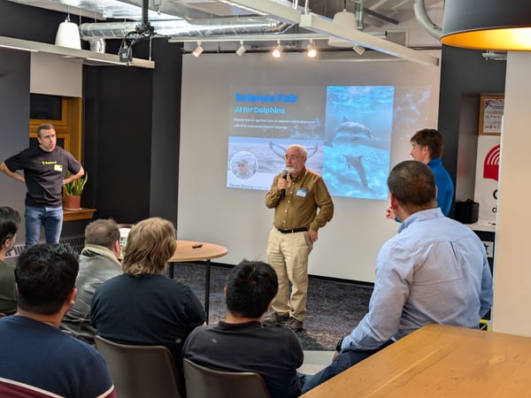 A photograph captures an indoor presentation where a speaker addresses an audience in front of a projection screen displaying a slide titled "AI for Dolphins."