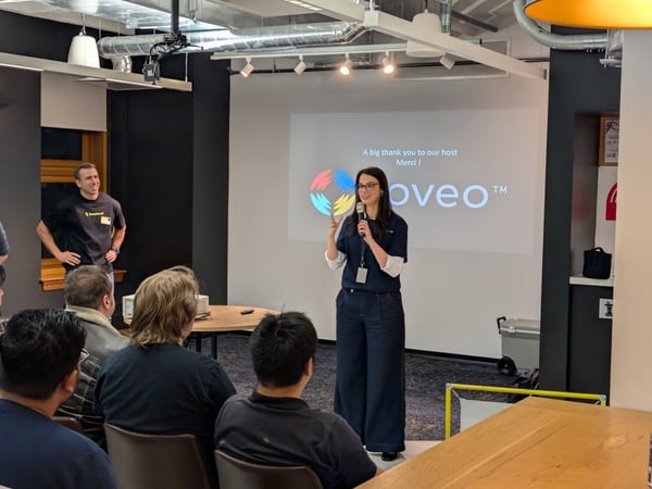 A photograph capturing an indoor presentation where a woman, holding a microphone, addresses a seated audience in front of a projection screen displaying a thank you message and the 'oveo' logo.