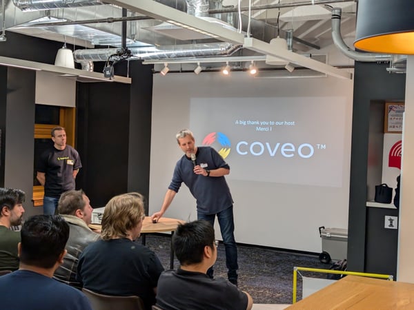 A photograph taken indoors showing a speaker presenting to an audience in a modern office or event space, with the Coveo logo and a thank you message projected onto a screen behind him.