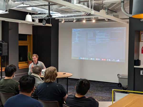 A man is giving a technical presentation or talk, holding a microphone while standing in front of a projector screen displaying lines of code and text to a small audience.