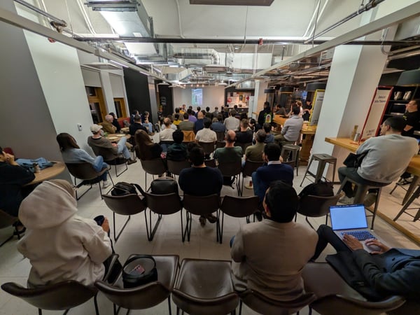This is a wide-angle photograph capturing a large audience attending an indoor presentation or seminar, seated in rows of chairs facing a projected screen at the front of a modern, industrial-style venue. Most attendees are visible from the back, with exposed ductwork visible on the ceiling.