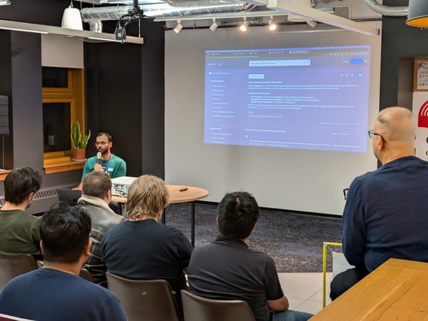 A photograph capturing a technical presentation indoors, showing a speaker holding a microphone next to a large projector screen displaying software documentation, while several audience members sit facing the front.