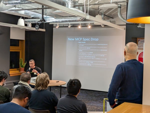 A man gives a technical presentation to a seated audience in a modern office space with an industrial ceiling, projecting a slide titled 'New MCP Spec Drop' onto a large screen.