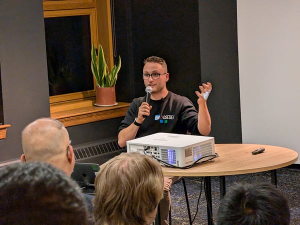 A man wearing glasses and a dark sweatshirt is giving a presentation, speaking into a microphone while standing behind a round wooden table holding a projector. The foreground shows the backs of several audience members' heads in a dimly lit indoor venue.