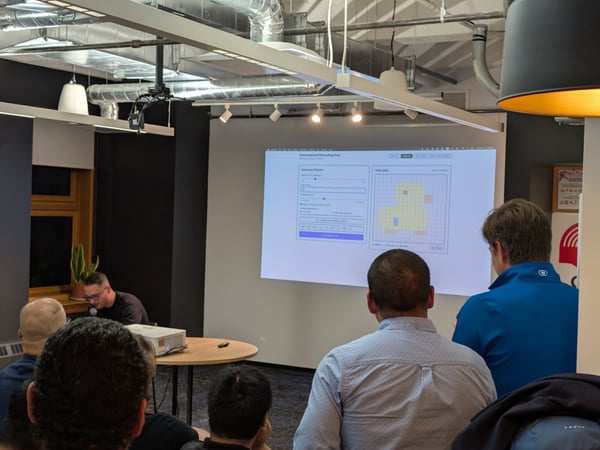 A photograph taken indoors during a technical presentation, showing several audience members viewing a projected screen that displays a computer interface featuring a grid with a yellow duck shape.