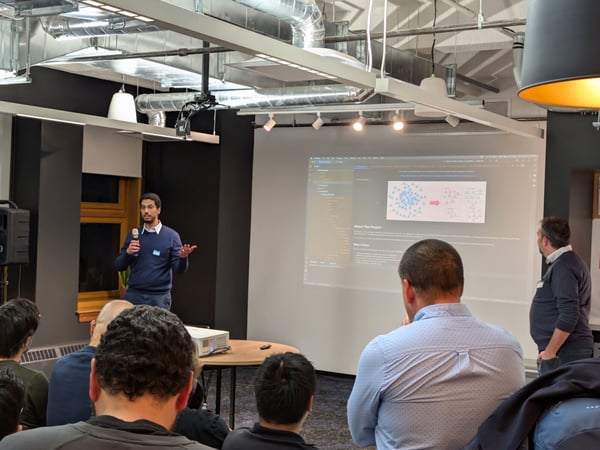 A photograph captures a man giving a presentation to an audience in a modern indoor venue with industrial-style ceilings, while a technical slide is projected onto a large screen behind him.