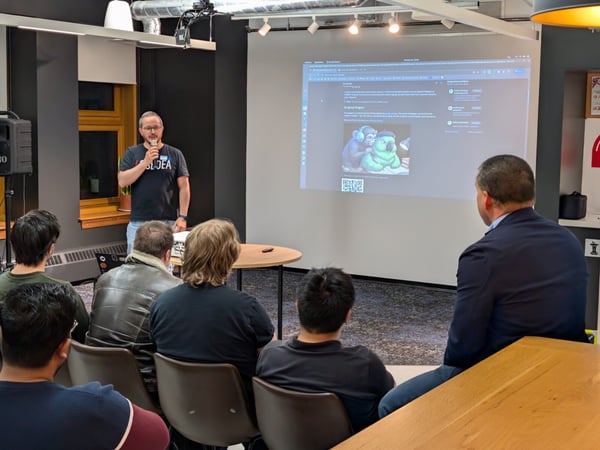 A photograph capturing an indoor presentation where a man wearing an "I ❤️ IDEA" t-shirt speaks into a microphone while standing next to a large screen displaying digital content, addressing a seated audience viewed mostly from the back.