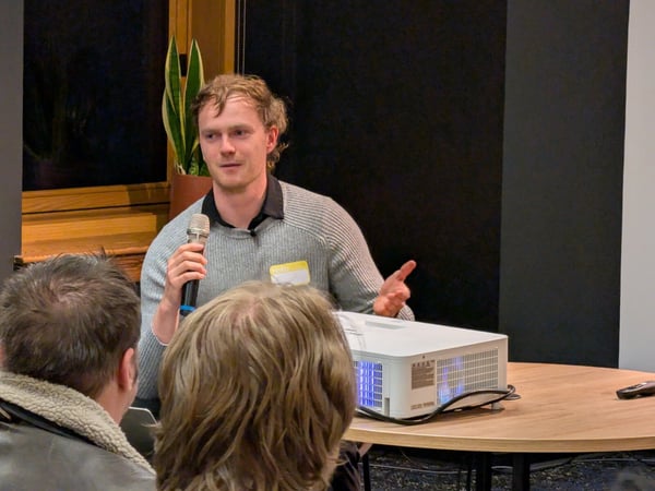A man wearing a grey sweater and a 'hello' name tag is speaking into a microphone during an indoor event, with the backs of two audience members visible in the foreground.