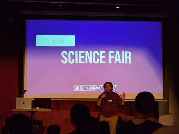 A man stands on a stage giving a presentation in front of a large screen that displays 'SCIENCE FAIR'.