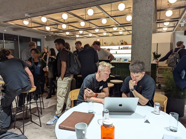 A candid photo capturing a busy indoor social or networking event, showing a large group of people mingling in a modern space with concrete ceilings and distinctive globe lighting fixtures. In the foreground, two men are seated at a white table, intently viewing content on a silver laptop.