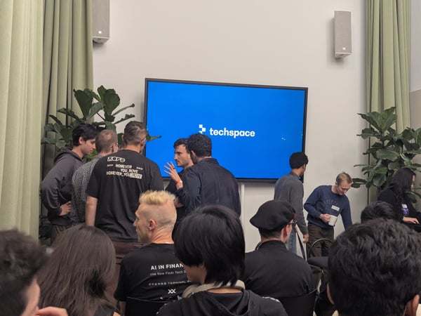 A candid photograph captures an indoor tech event or meetup, showing a crowd of people mingling and engaging in discussions, primarily wearing dark clothing.