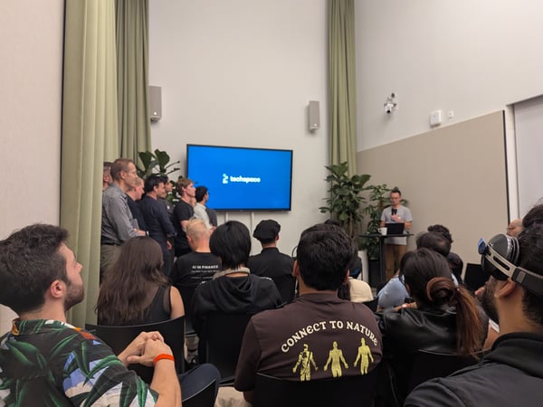 A photograph capturing an indoor presentation or tech talk, showing a speaker at a podium addressing a large audience, with others standing nearby and a screen displaying the "techspace" logo.