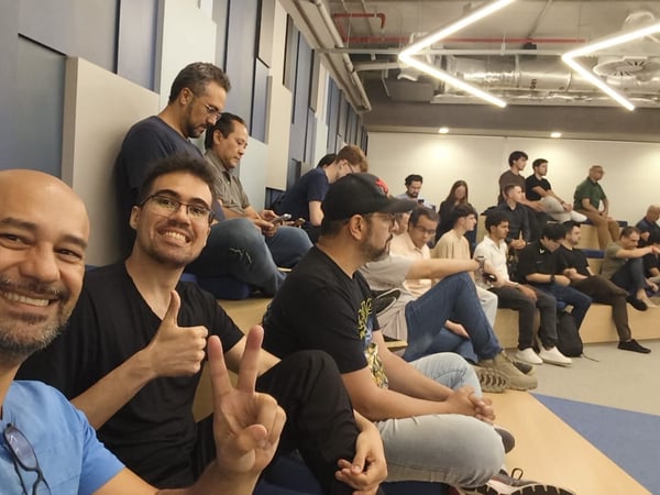 A candid photo taken from the front row of a tiered seating area, showing a large group of people attending an indoor event or presentation. Two men in the immediate foreground are smiling and posing for the camera with a thumbs up and a peace sign.