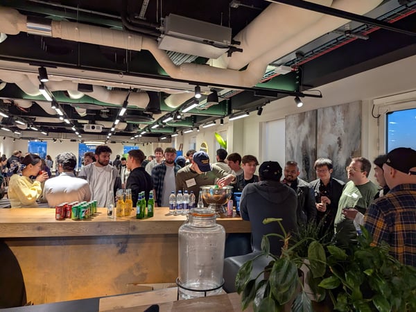 A candid photograph captures a large group of people socializing and networking around a wooden bar counter in a modern indoor space with exposed ceiling ductwork and track lighting.
