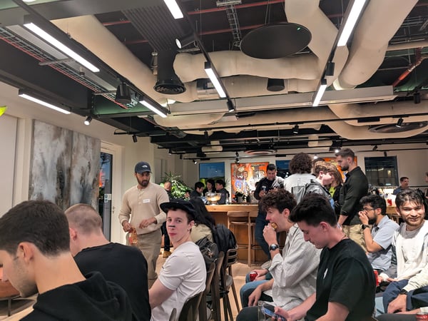 A candid photograph capturing a social or networking event held indoors, showing a large group of people gathered in a modern space with exposed industrial ceiling ductwork and bright strip lighting. Several attendees are seated in the foreground, while others mingle near a bar area in the background.