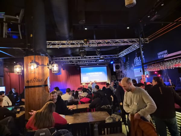 A crowded indoor social event at a bar or venue, with a presentation screen in the background displaying 'AI TINKERERS FÉVRIER 2024'.