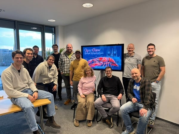 A group of twelve people posing for a group photograph in an office setting, positioned in front of a screen displaying 'OpenClaw Global Unhackathon'.