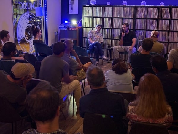 An indoor event showing two men speaking to a seated audience in front of shelves filled with vinyl records.