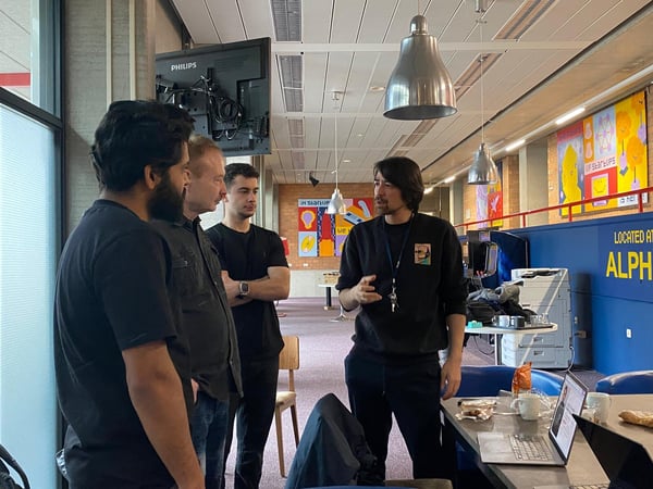 A group of four men are engaged in a conversation within a modern coworking or office space, with one man gesturing while speaking near an open laptop.