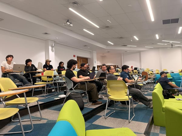 A group of people sitting in a tiered classroom or lecture hall, most of whom are using laptops and looking toward the front of the room.