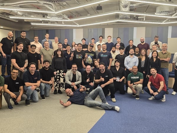 A large group of approximately 38 colleagues poses for a group photograph in a modern, well-lit office space with blue and gray paneled walls. The team is arranged in multiple rows, with several individuals kneeling or squatting in the foreground and one person lying down playfully.