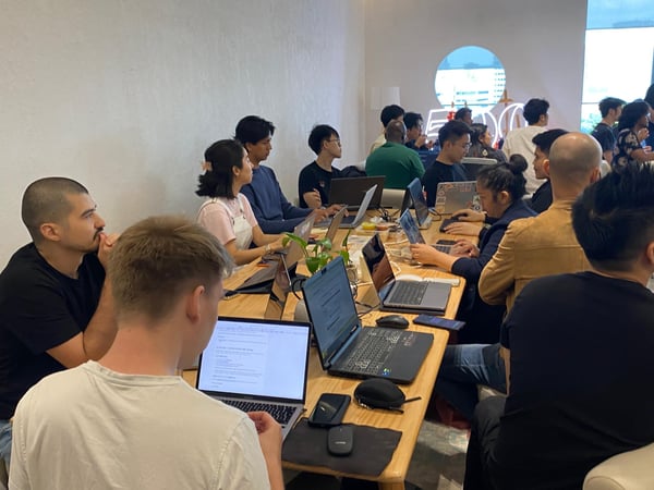 A group of people are working on their laptops at long wooden tables in a bright, modern indoor space, likely a co-working office.