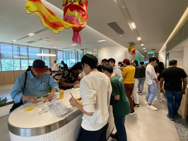 A group of people are gathered in a modern office pantry or common area, some eating at a counter, with traditional Chinese lion dance heads hanging from the ceiling.