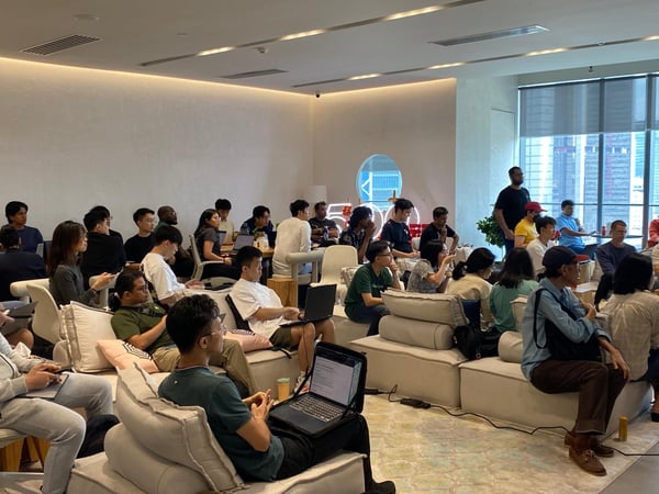 A group of people are seated in a modern, well-lit indoor space, many using laptops and appearing to attend a presentation or meeting.