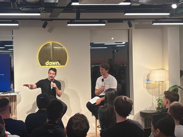 A photograph capturing an indoor panel discussion or Q&A session, featuring two men seated on stools addressing a large, dimly lit audience. Behind the speakers, a large television screen displays a 'Check Device Power' error message, and a glowing sign on the wall reads 'dawn.'.