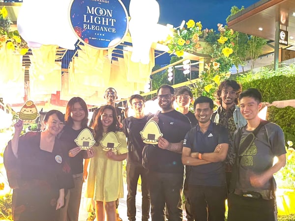 A group of nine individuals is posing for a photo outdoors at night under large, illuminated white lanterns and festive yellow decorations, likely celebrating the Mid-Autumn Festival. A circular sign advertising 'MOON LIGHT ELEGANCE' is visible above them, and several people are holding small, cartoon onigiri-shaped cutouts.
