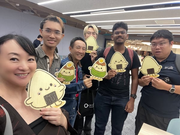 A group selfie featuring six people (five men and one woman) smiling and posing indoors while holding up large, cartoon cutout illustrations of anthropomorphic onigiri (rice balls). The setting appears to be a modern indoor event or office space.