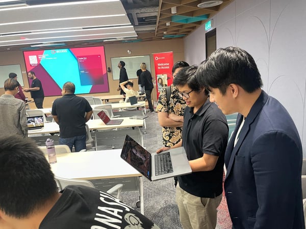 A candid photograph taken indoors showing a group of people attending a technical workshop or meeting, with two men in the foreground intently examining content on a laptop screen.