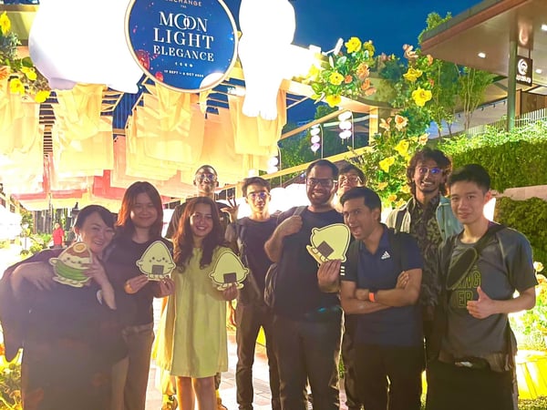 A group of nine people poses for a photo outdoors at night amidst festive decorations, including large illuminated white rabbit lanterns and a sign advertising the 'Moon Light Elegance' event.