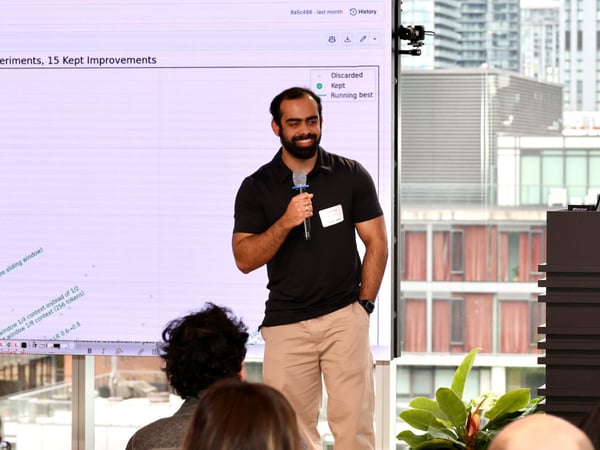 A man stands in front of a large screen giving a presentation, holding a microphone and smiling while an audience sits in the foreground.