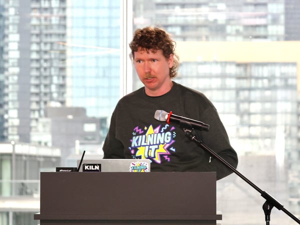 A man with curly hair and a mustache is standing behind a podium, giving a presentation with a large window and cityscape in the background.