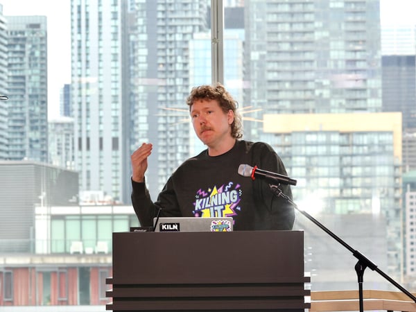 A man with a mustache and curly hair is giving a presentation behind a podium, with a dense city skyline visible through the window behind him.
