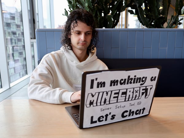 A man with curly hair wearing a white hoodie sits at a table, working on a laptop that has a sign on its back inviting conversation about a Minecraft server setup tool.