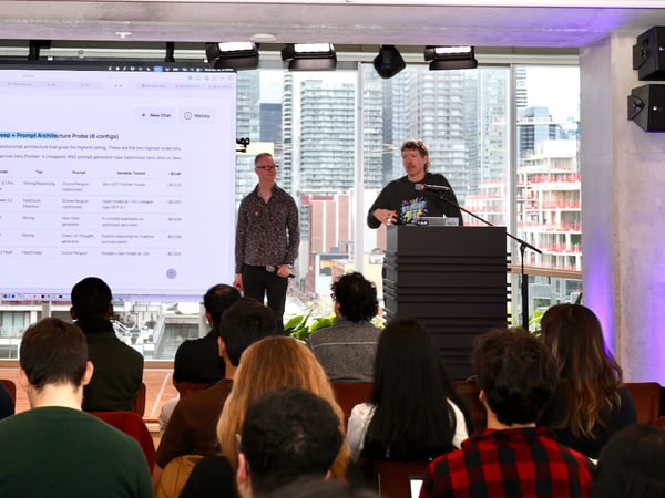 A man stands at a podium giving a presentation to an audience, with a large screen displaying a data table about AI model performance.