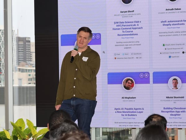A man in an olive green cardigan is speaking into a microphone while standing in front of a large screen displaying project information during a presentation.