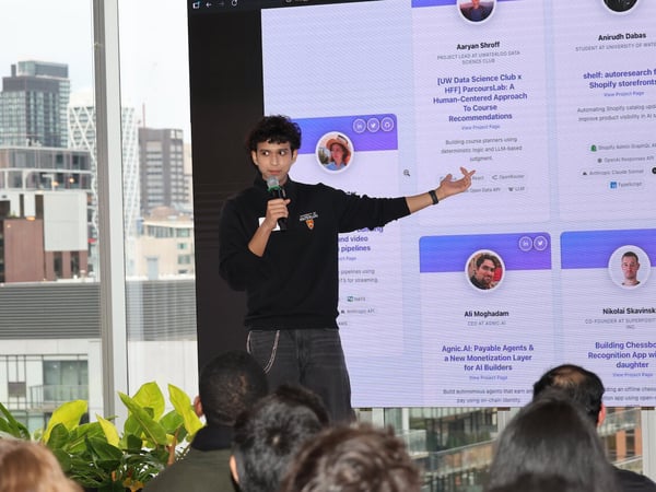 A young man is giving a presentation in front of a large screen displaying project profiles, with a city skyline visible through a window in the background.
