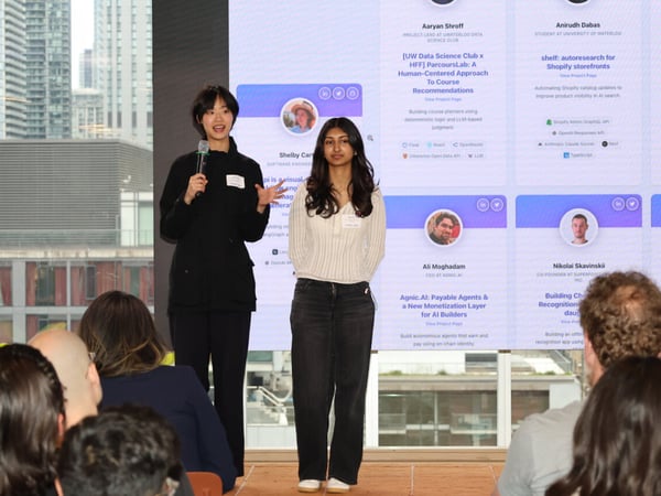 Two women are giving a presentation on a stage in front of a large digital screen displaying project profiles, with an audience in the foreground and a city skyline visible through windows.