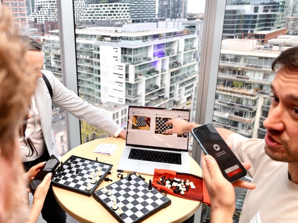 A group of people are gathered around a small table in a high-rise building, demonstrating a chess-related application on a laptop and smartphones.