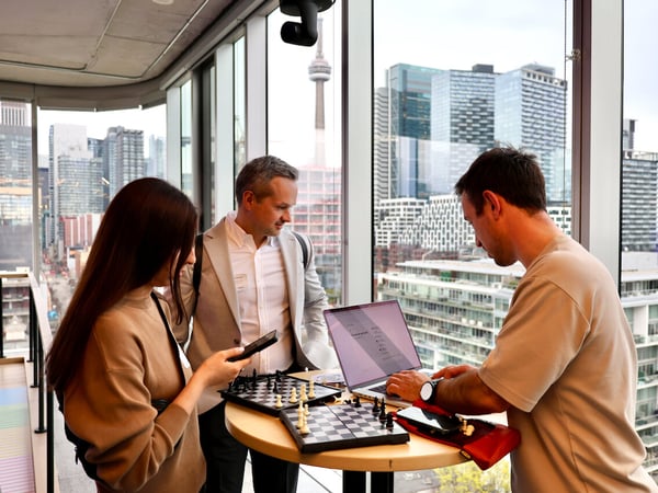 Three people are gathered around a high table with a laptop and two chessboards in a room with floor-to-ceiling windows overlooking the Toronto skyline.