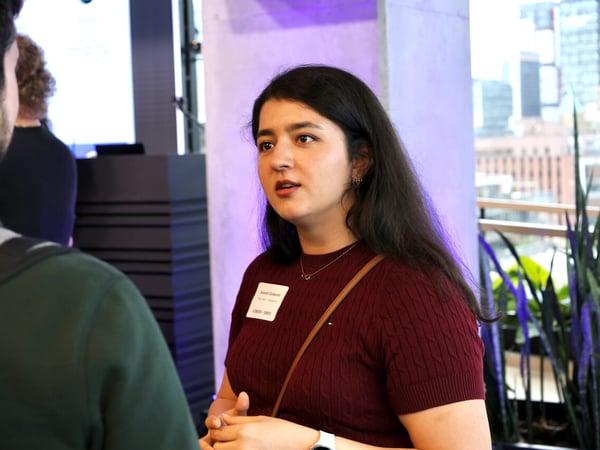 A woman in a maroon top is engaged in a conversation with a man at a professional networking event, with a city skyline visible in the background.