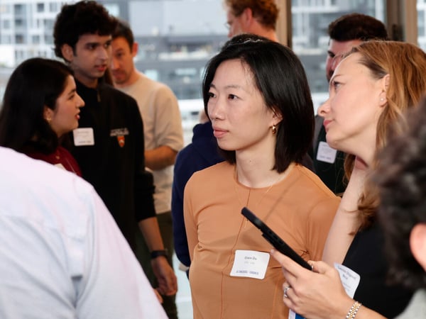 A group of people are gathered at a professional networking event, with a woman in a tan top as the central focus.