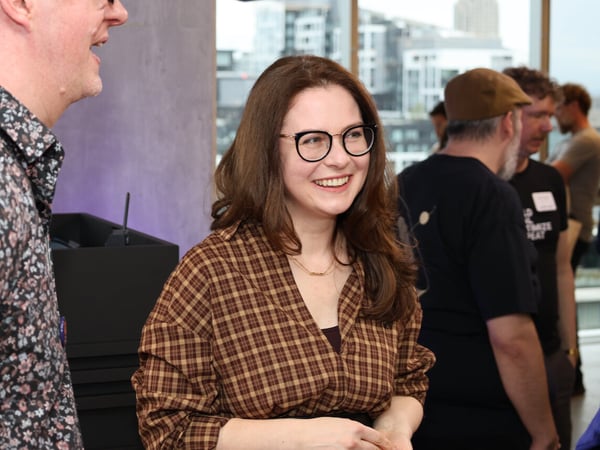 A woman with glasses and a checkered shirt smiles during a social gathering in an indoor space with a city view.