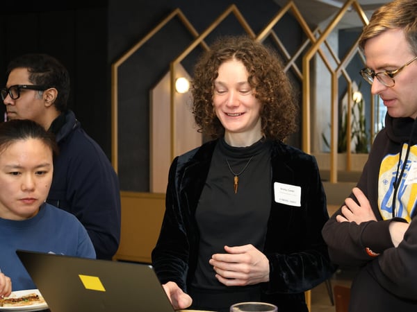 A group of people are gathered around a laptop at an indoor event, with a woman in a black velvet blazer smiling at the screen.
